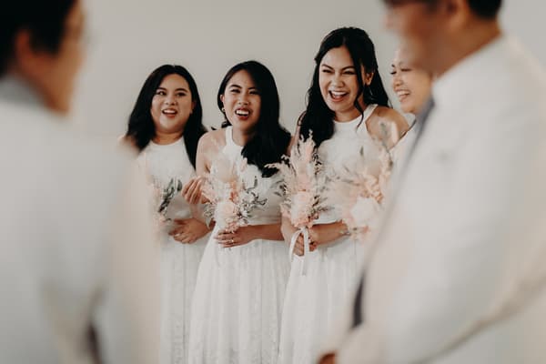 Three bridesmaids in white dresses holding bouquets stand together smiling, with two men in white suits partially visible in the foreground at Royal on the Park.