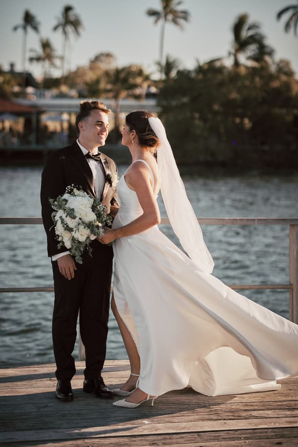 The bride Ashleigh in a white wedding gown and veil stands facing the groom James in a black tuxedo holding a bouquet of white flowers on a wooden deck by the water at Sandstone Point Hotel.
