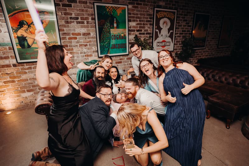 A group of wedding guests pose energetically on the floor at the Sandstone Point Hotel — Cellar during the reception stage, with brick walls and vintage posters in the background.