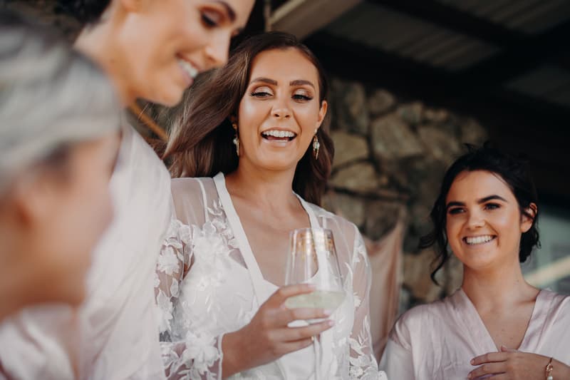 The bride Chelsea in a white lace robe holding a glass, smiling and talking with two bridesmaids in light pink robes at Preston Peak Winery Functions Centre.