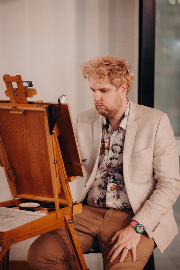 The groom, Adam, sits at an easel painting or drawing during the reception at Ocean View Estates — Function Room.