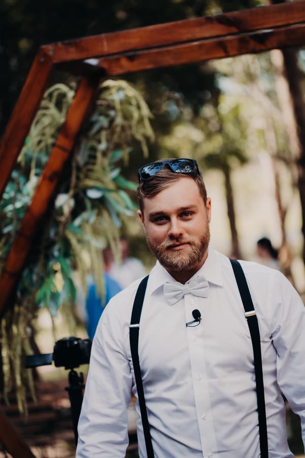 The groom Trent stands near a wooden arch decorated with greenery at Ocean View Estates, wearing a white shirt, bow tie, black suspenders, and sunglasses on his head.