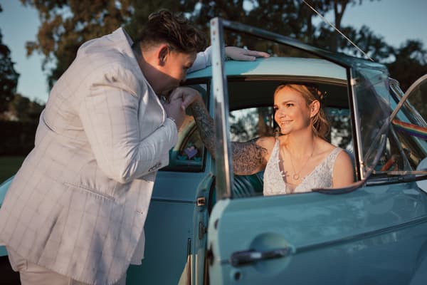 Mandy, the bride in a white lace dress, sits inside a vintage light blue car while Selina, the bride in a light-colored checked suit, leans in through the open door and kisses Mandy's hand. Cinematic videography wedding by She Said Yes.
