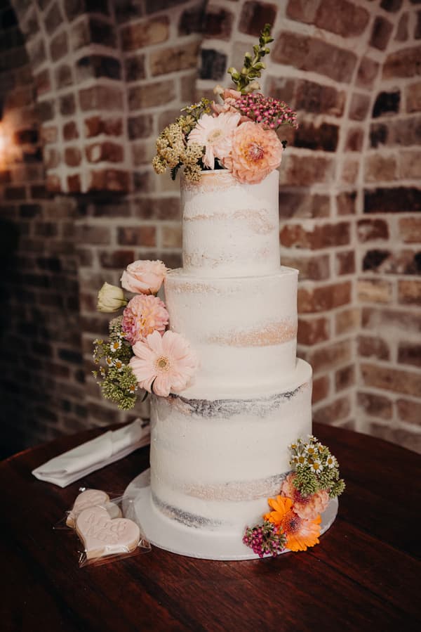 Three-tier white wedding cake decorated with assorted fresh flowers on a wooden table at Sandstone Point Hotel — Cellar, accompanied by heart-shaped cookies wrapped in plastic.