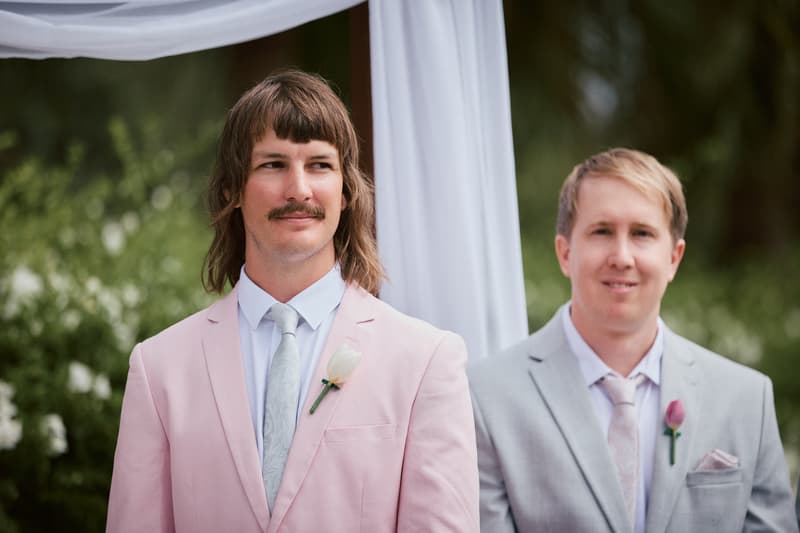 Two men stand side by side at the ceremony stage at Eatons Hill Hotel — Lakeside, both wearing suits with boutonnieres; the man on the left wears a pink suit with a white rose boutonniere, and the man on the right wears a light gray suit with a pink rose boutonniere.