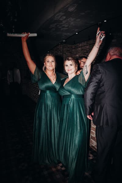 Two bridesmaids in matching green dresses raise their arms in celebration at the reception stage of, with other guests partially visible nearby. ABIA Awards wedding photography by She Said Yes.