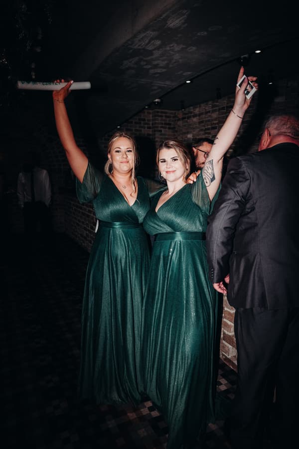 Two bridesmaids in matching green dresses raise their arms in celebration at the reception stage of Sandstone Point Hotel — Cellar, with other guests partially visible nearby.