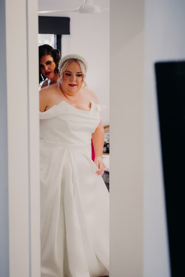 The bride Jacquelyne is being assisted by a bridesmaid while getting ready indoors.