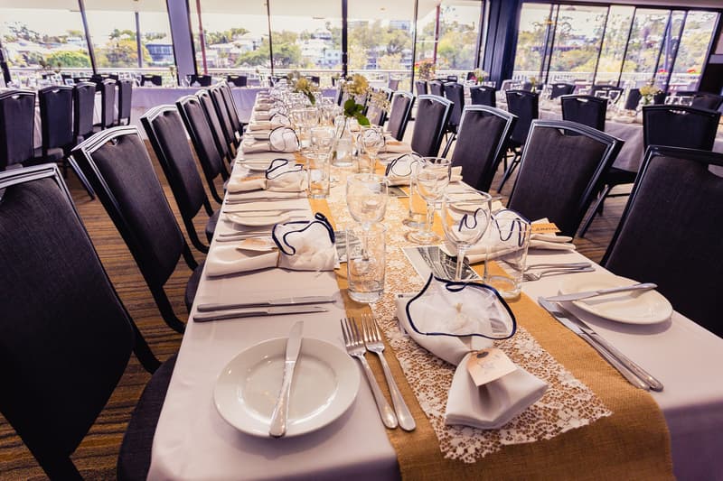 Long dining table set for a wedding reception with white tablecloth, lace and burlap runner, neatly arranged plates, cutlery, glasses, and napkins with name tags at Toowong Rowing Club — The Malouf Room.