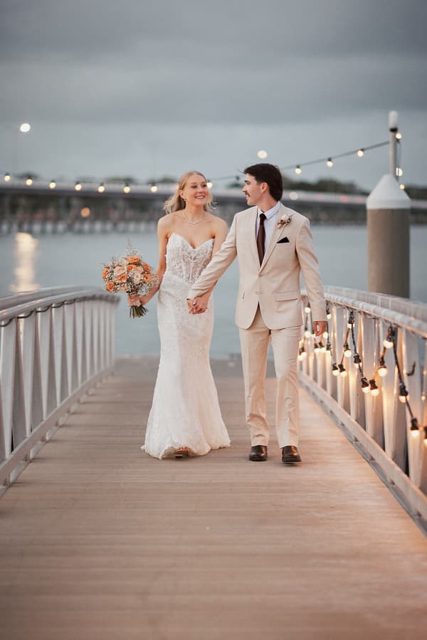 Morgan and Joel walk hand in hand on a wooden pier at Sandstone Point Hotel, with Morgan holding a bouquet of flowers and string lights decorating the railing.