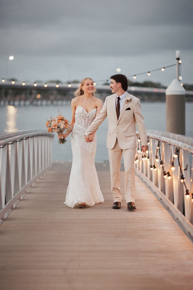 Morgan and Joel walk hand in hand on a wooden pier, with Morgan holding a bouquet of flowers and string lights decorating the railing. ABIA Awards wedding photography by She Said Yes.