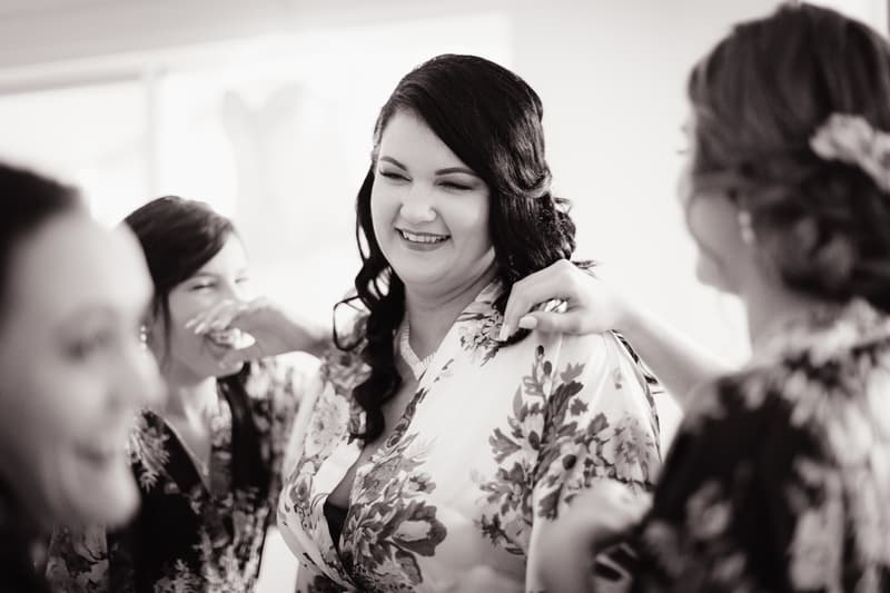 The bride Kelly is smiling and surrounded by bridesmaids helping her with her floral robe during bridal preparations at Novotel Sunshine Coast Resort.