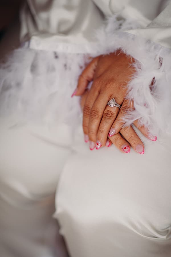 Close-up of the bride's hands resting on her lap, showing a pear-shaped engagement ring and pink nail art with heart designs, wearing a white garment with feathered cuffs.