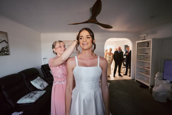 The bride Ashleigh stands in a room wearing her wedding dress while a woman in a pink dress adjusts her hair. In the background, three guests stand in a doorway watching.