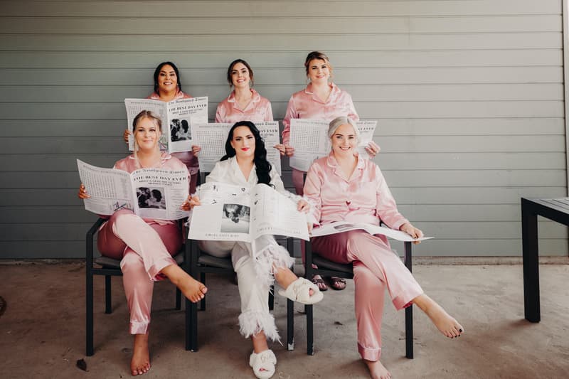 The bride Mindy in white pajamas and four bridesmaids in matching pink pajamas sit on chairs holding newspapers at Sandstone Point Hotel.