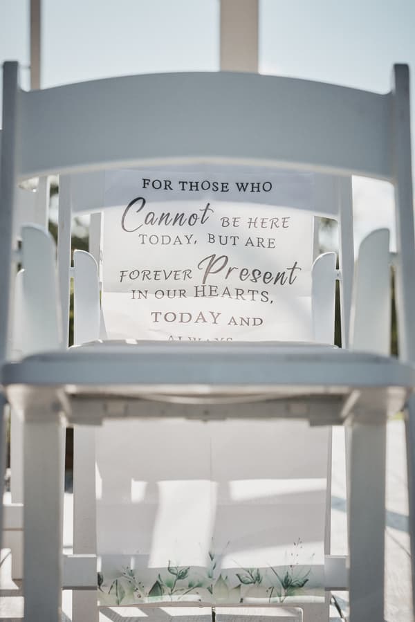 A white chair at Sandstone Point Hotel — The Pavilion with a sign that reads 'For those who cannot be here today, but are forever present in our hearts, today and always.'