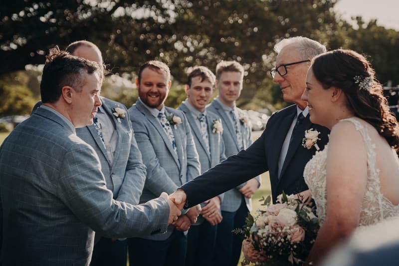 The bride's father shakes hands with the groom at Sandstone Point Hotel — Rustic Arbour during the ceremony, with the bride holding a bouquet and groomsmen standing in the background.