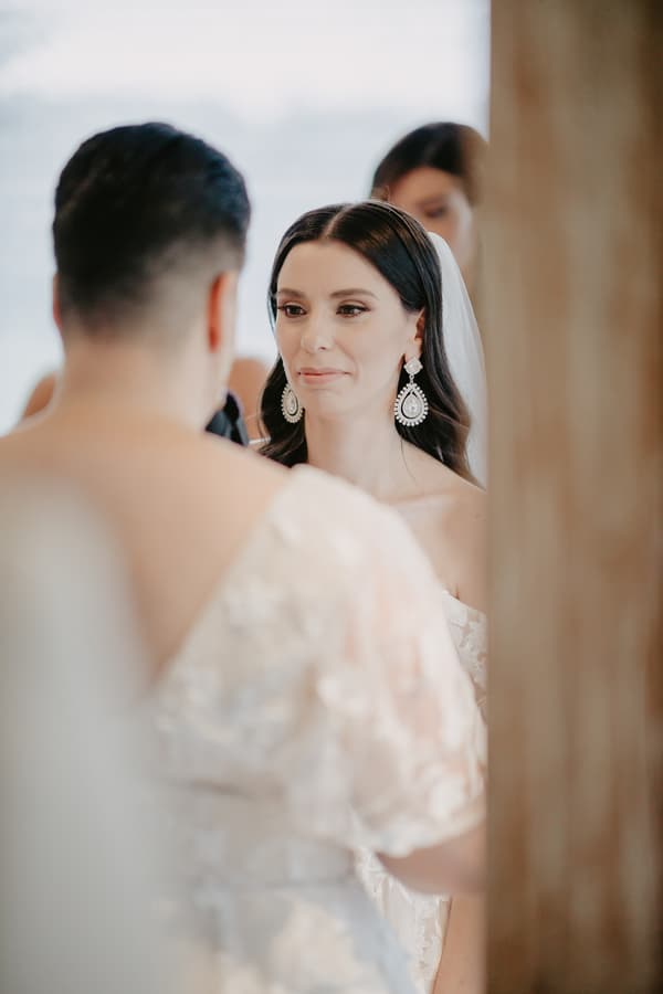 The bride with long dark hair and large earrings faces another woman in a white dress, with a blurred figure in the background.