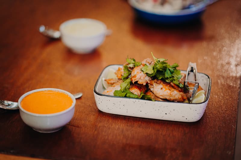 A serving dish with grilled chicken garnished with fresh herbs and lime wedges on a wooden table at Yabbaloumba Retreat — The Shed, accompanied by two small bowls of sauces.