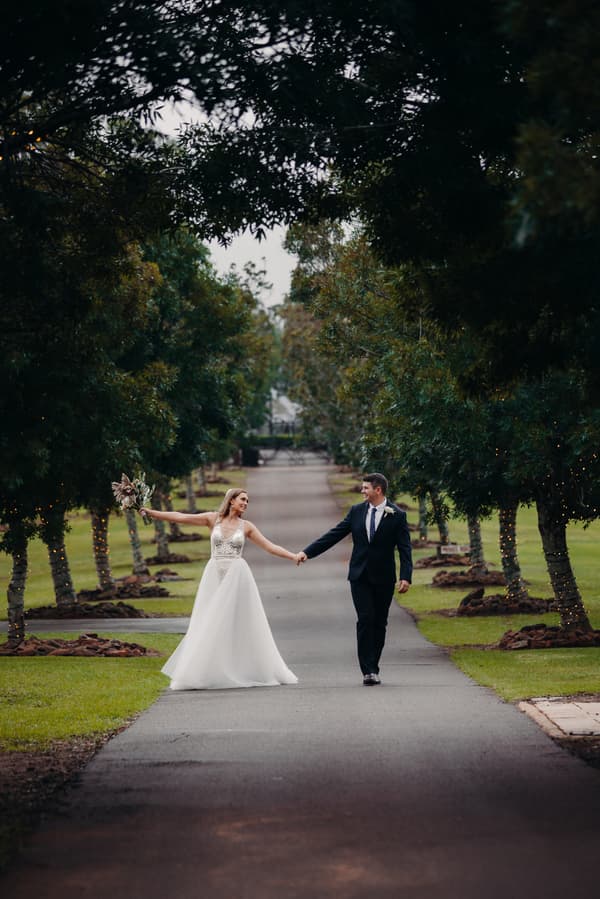 Chelsea the bride and Brent the groom walk hand in hand down a tree-lined path at Preston Peak Winery Functions Centre, with Chelsea holding a bouquet.