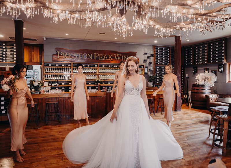 The bride Chelsea in a white wedding gown stands in the center of Preston Peak Winery Functions Centre with four bridesmaids in matching blush dresses around her, inside a room with wooden floors, wine racks, and a chandelier.