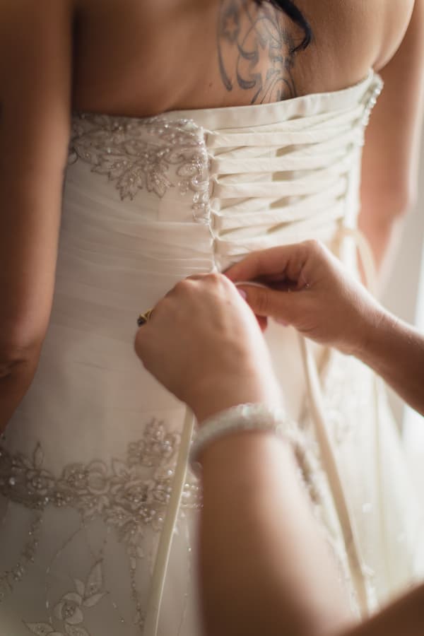 The bride is having the back of her wedding dress laced up by another person.