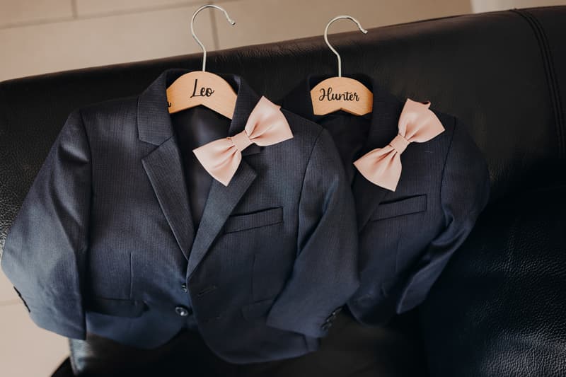Two dark gray children's suit jackets with pale pink bow ties on wooden hangers labeled 'Leo' and 'Hunter' placed on a black leather couch.