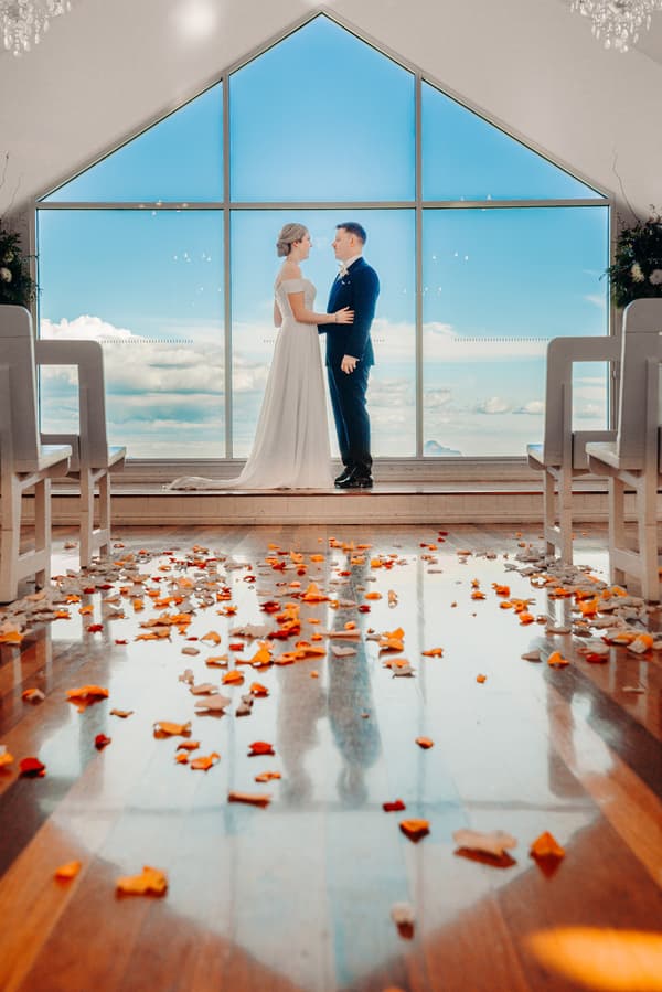 Courtney and Liam stand facing each other inside Tiffany's Maleny chapel with a large triangular window showing a blue sky and clouds behind them. The wooden floor is scattered with orange and white flower petals.