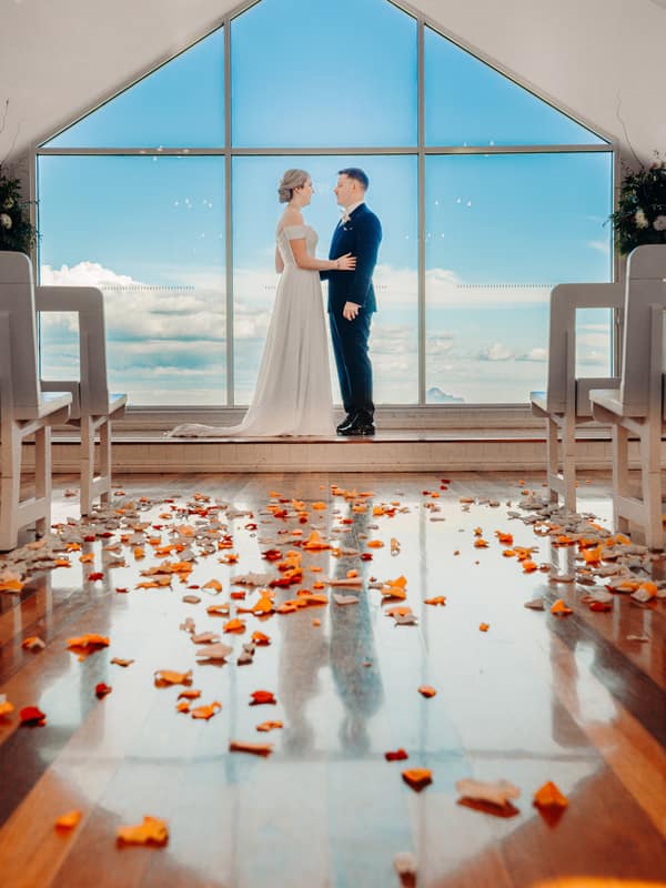 Courtney and Liam stand facing each other inside Tiffany's Maleny chapel with a large triangular window showing a blue sky and clouds behind them. The wooden floor is scattered with orange and white flower petals.