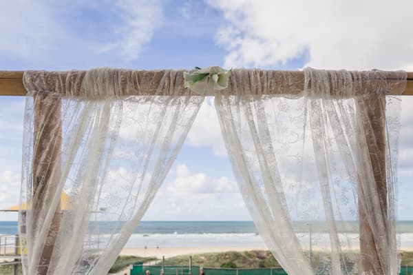 Decorative lace fabric draped over a wooden wedding arch at Bilinga Beach Weddings — The Terrace, with the beach and ocean visible in the background under a partly cloudy sky.