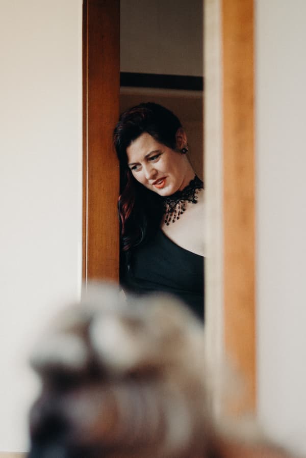 A woman in a black dress and black beaded necklace is partially visible behind a wooden door frame, looking downward.