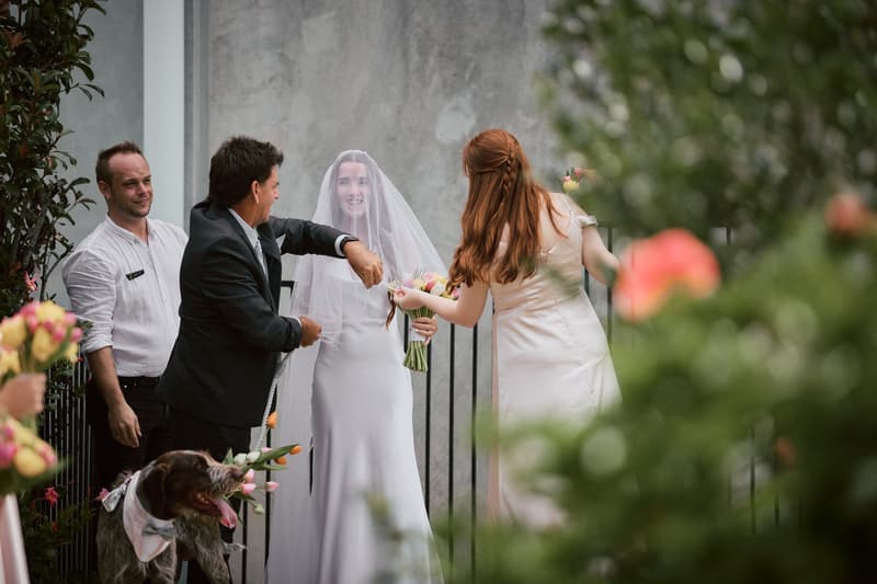 The bride Olivia, wearing a veil and holding a bouquet, stands near a man in a suit and a woman in a light dress at Eatons Hill Hotel — Lakeside. A man in a white shirt and a dog with a bandana are also present nearby.