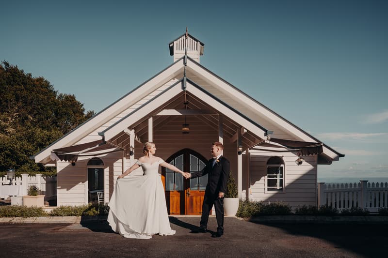 Courtney the bride and Liam the groom hold hands and pose in front of a white chapel building at Tiffany's Maleny during their couple portraits session.
