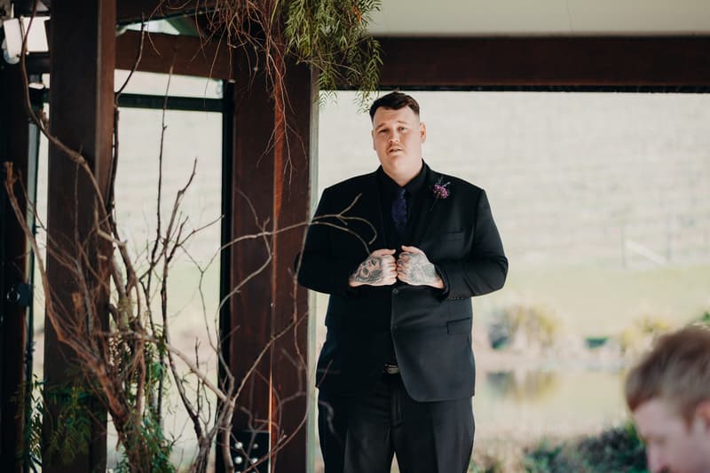 Adam, the groom, stands alone on the ceremony stage at Ocean View Estates — On The Lake, wearing a black suit with a purple boutonniere and tie.