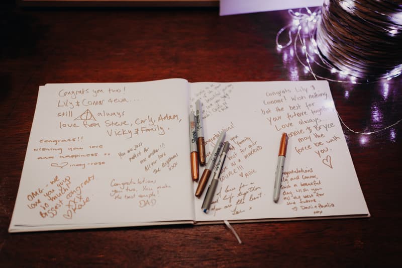 Open wedding guestbook with handwritten congratulatory messages for Lilly and Connor, placed on a wooden table at Yabbaloumba Retreat — The Shed, with several metallic pens and string lights nearby.