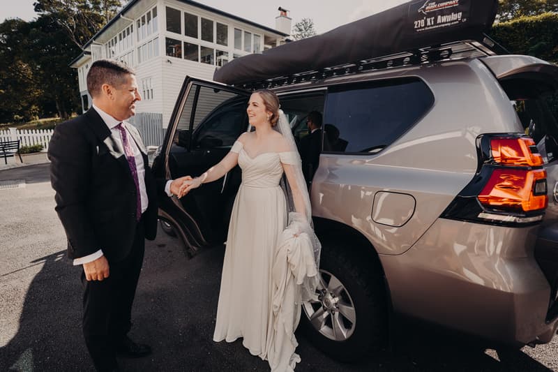 The bride Courtney, wearing a white wedding dress and veil, holds hands with an older man in a suit and purple tie outside a vehicle at Tiffany's Maleny — Chapel.