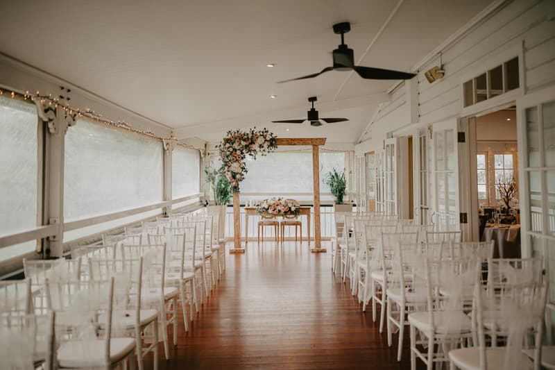 Empty indoor wedding ceremony setup with rows of white chairs facing a wooden arch decorated with flowers and greenery, with a small table underneath the arch.