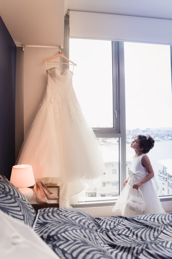 A flower girl in a white dress and tiara looks up at a hanging wedding gown beside a window with a cityscape view at Hillstone St Lucia.