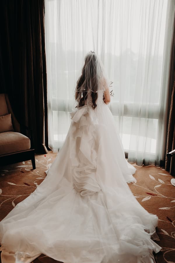 The bride stands alone facing a large window with sheer curtains at Royal on the Park, wearing a long white wedding gown with a veil and holding a bouquet.
