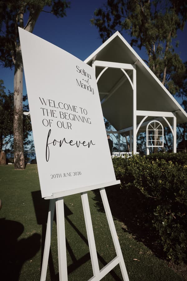 A white welcome sign for Selina and Mandy's wedding dated 20th June 2026 is displayed on an easel outside The Pavilion at Sandstone Point Hotel, with the white ceremony structure and greenery visible in the background.