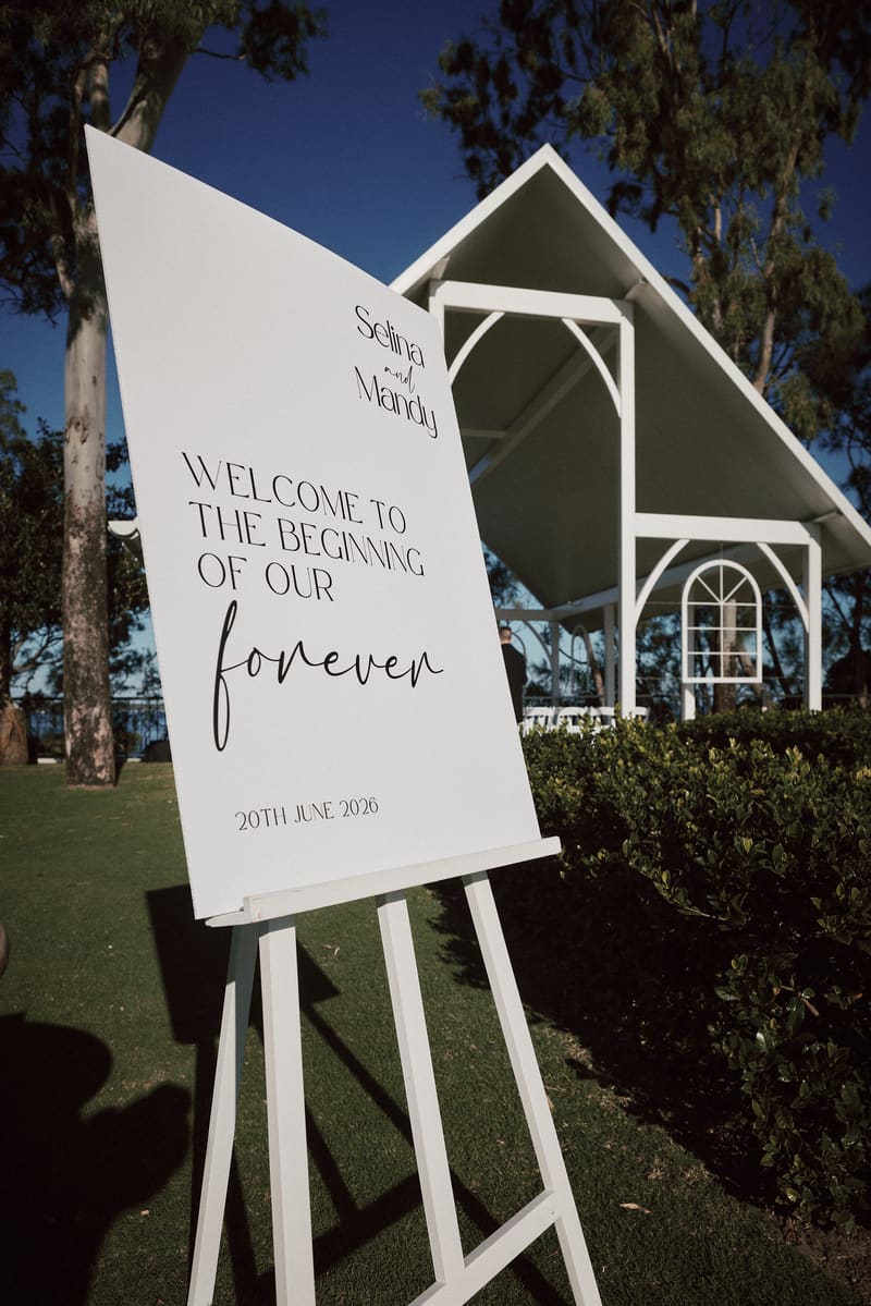 A white welcome sign for Selina and Mandy's wedding dated 20th June 2026 is displayed on an easel outside The Pavilion, with the white ceremony structure and greenery visible in the background. Cinematic wedding videographer Gold Coast by She Said Yes.