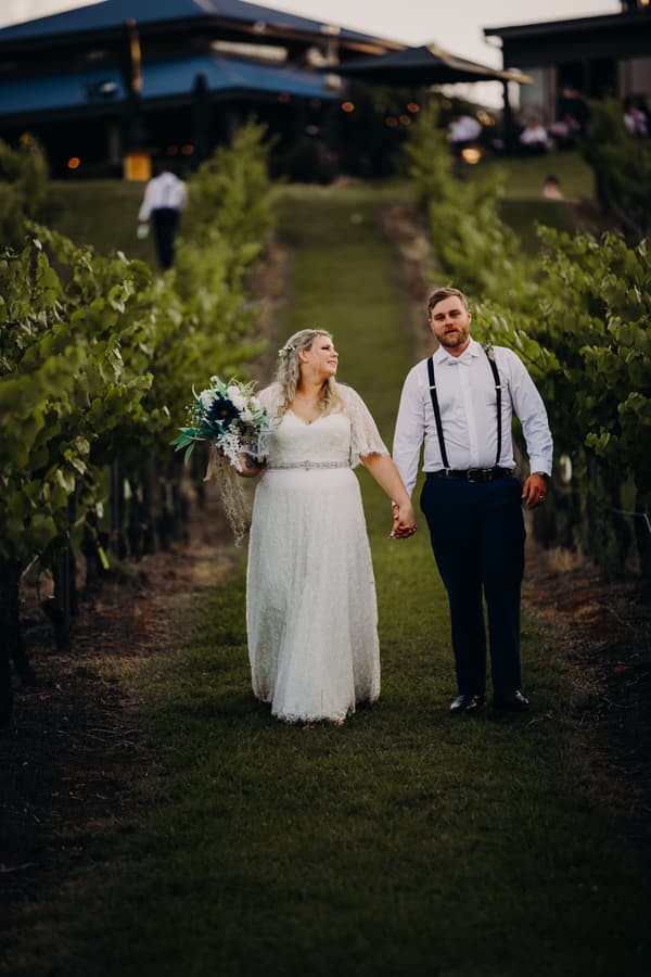 Annie in a white wedding dress holding a bouquet and Trent in a white shirt with suspenders walk hand in hand down a grassy path between rows of grapevines at Ocean View Estates.