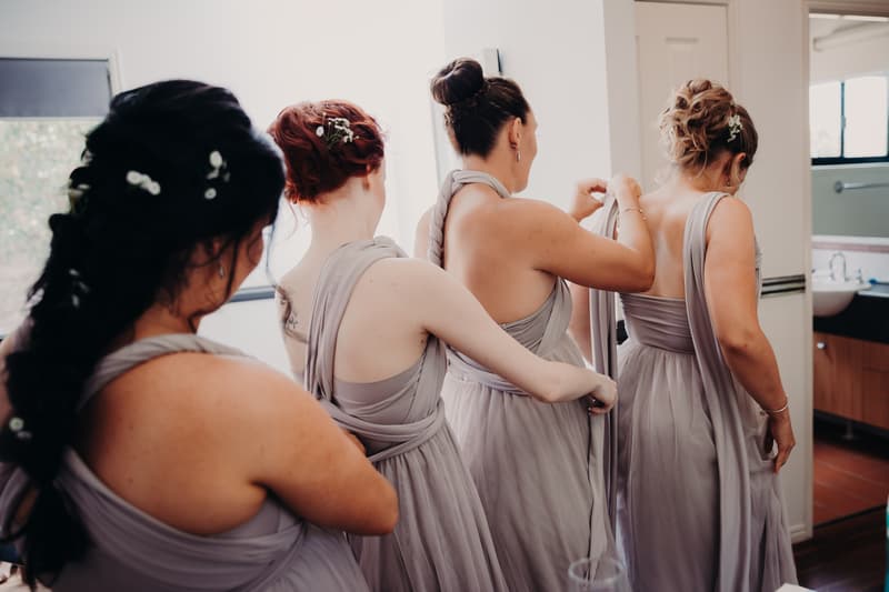 Four bridesmaids in matching lavender dresses are helping each other adjust their gowns in a room at Ocean View Estates.