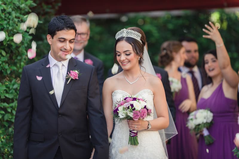 Pasha and Maryam walk together at Hillstone St Lucia — The Quartyard with bridesmaids in purple dresses throwing flower petals behind them.