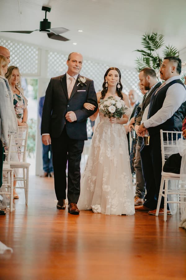 The bride walks down the aisle arm-in-arm with an older man, likely her father, while guests stand on either side watching.