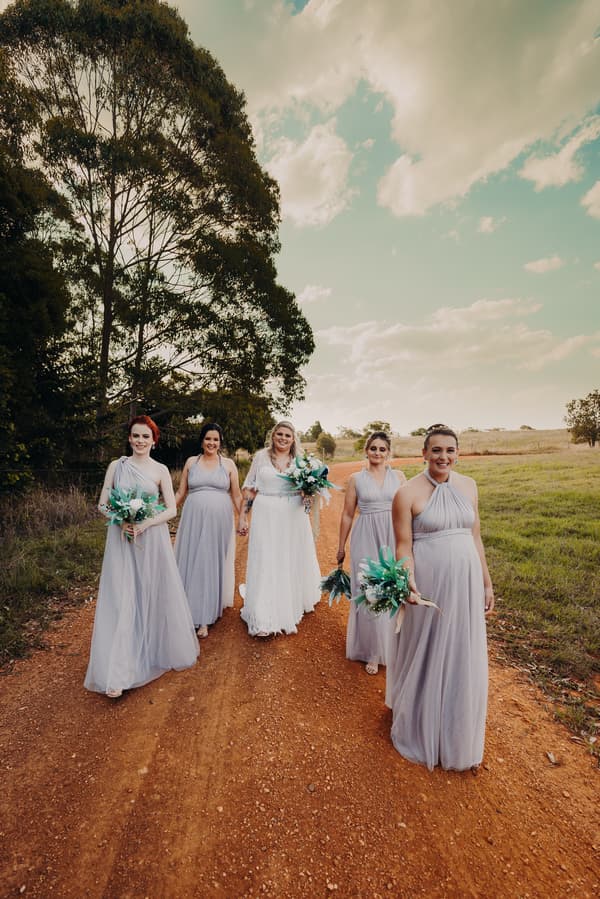The bride Annie walks along a dirt path at Ocean View Estates with four bridesmaids in matching lavender dresses, each holding bouquets.