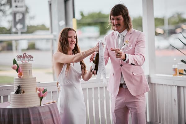 Olivia the bride and Jake the groom stand near a white railing at White Horse Ranch reception stage, with Olivia opening a champagne bottle and Jake holding two champagne flutes. A white tiered wedding cake decorated with pink flowers and a 'Mr & Mrs' topper is on a nearby table.