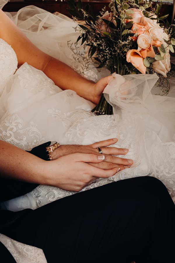 The bride and groom hold hands resting on the bride's lace wedding dress, with the bride holding a bouquet of peach and white roses and greenery.