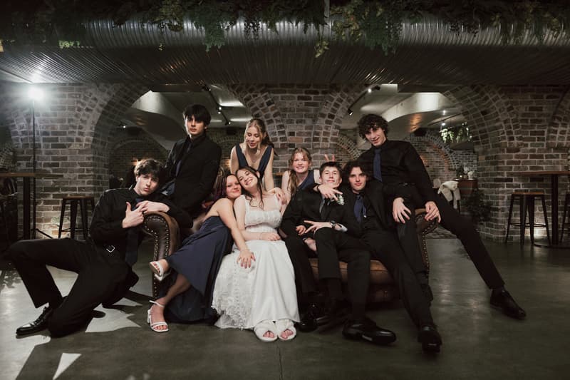Terion in her wedding dress sits on a couch surrounded by Tyler and seven other wedding party members dressed in black suits and navy dresses at Sandstone Point Hotel — The Cellar.