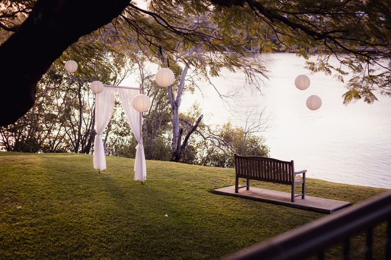 A grassy outdoor area at Toowong Rowing Club with a wooden bench on a concrete slab, a white fabric archway, and white paper lanterns hanging from tree branches near a body of water.